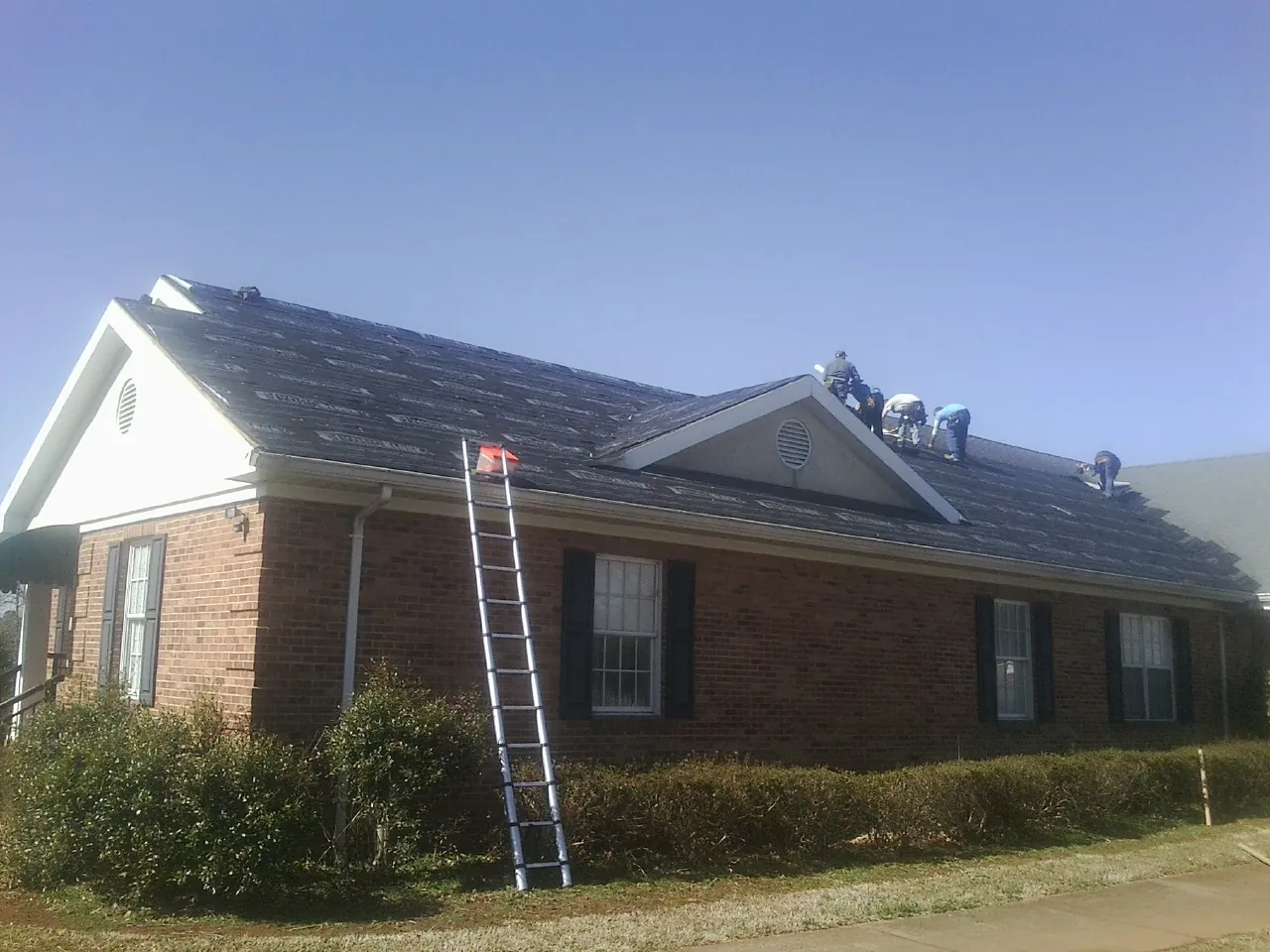 Crew tearing shingles off a brick ranch roof with a ladder set — Metal Roof Repair work by our crew in Hebron